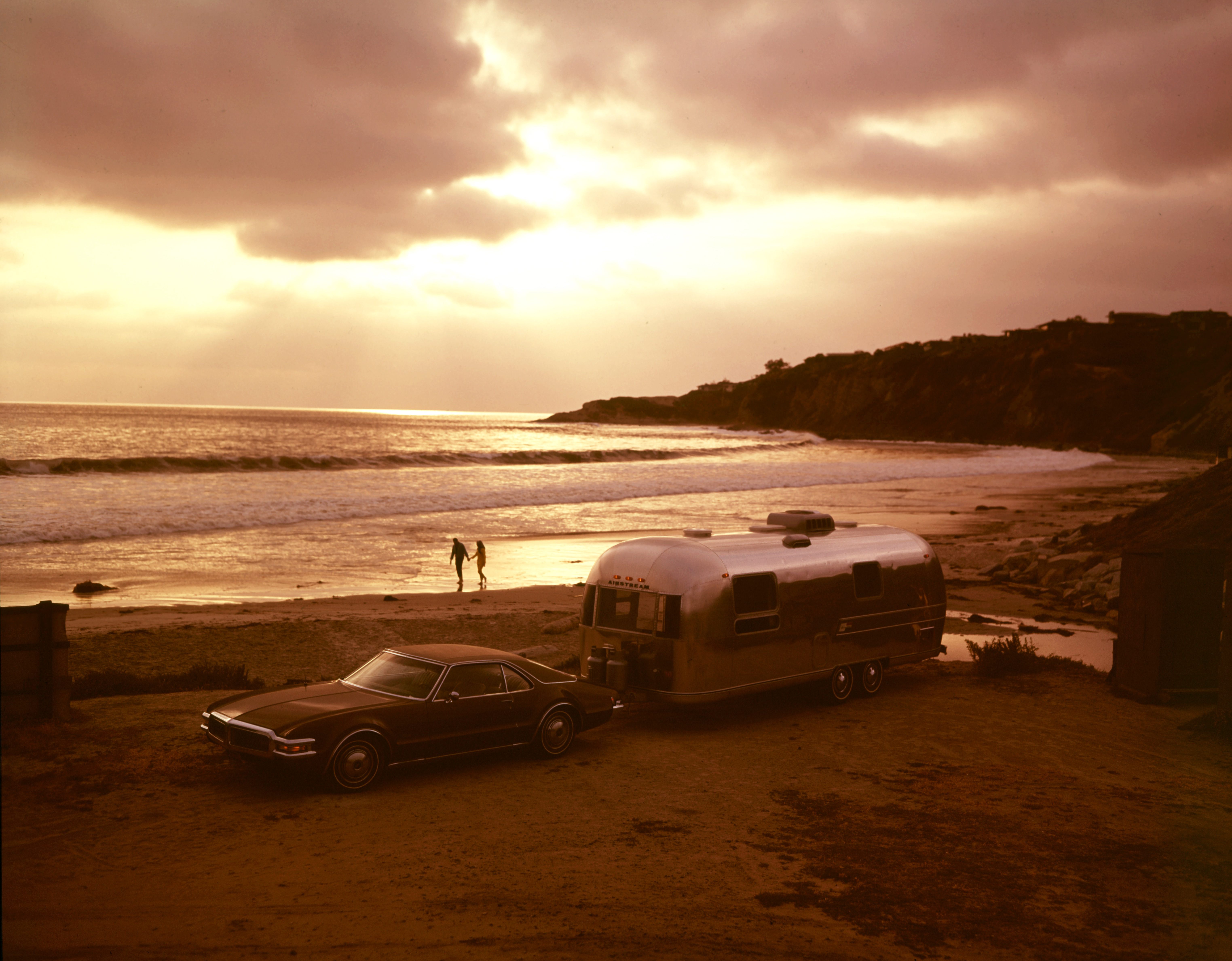 A vintage car towing a silver Airstream trailer is parked on a sandy beach at sunset, with two people walking along the shoreline and waves gently rolling in under a cloudy sky.