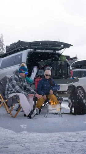 Two people in winter gear sit in chairs beside a small fire with a dog nearby, set against a snowy backdrop with parked cars and trees.