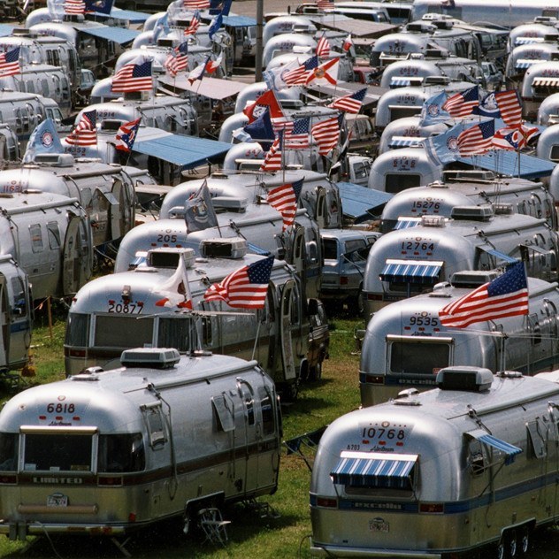 Numerous silver Airstream trailers are parked closely together on grass, each displaying American flags, creating a festive and patriotic atmosphere.