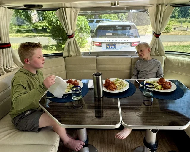 Two boys are seated at a dining table in a camper, enjoying plates of pasta and salad. A parked SUV is visible outside the window.