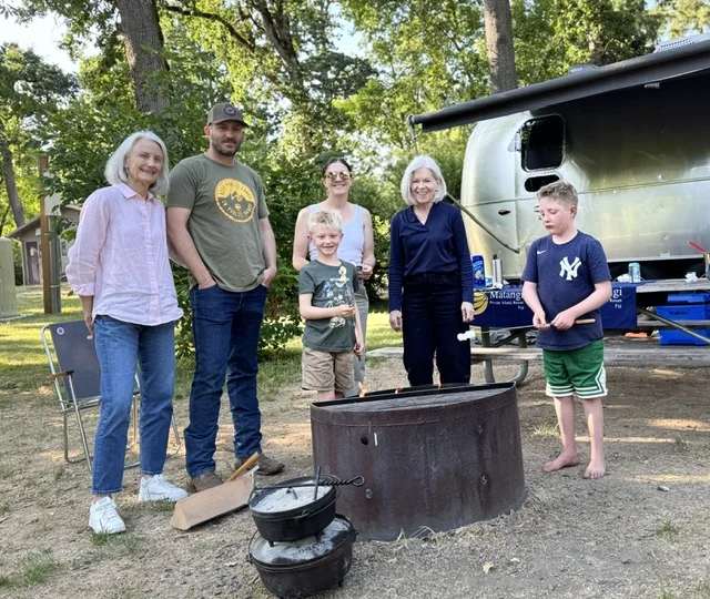A group of six people roasting marshmallows around a fire pit in a campsite with a trailer and trees in the background.