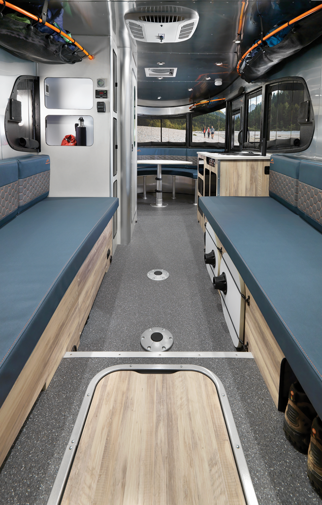 Interior of a modern RV with blue cushioned benches, wood accents, storage, and rear window view of a natural landscape.