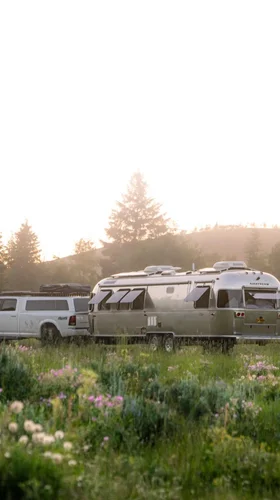 A white truck towing a silver Airstream trailer parked in a lush green field with wildflowers, surrounded by trees and hills at sunrise.