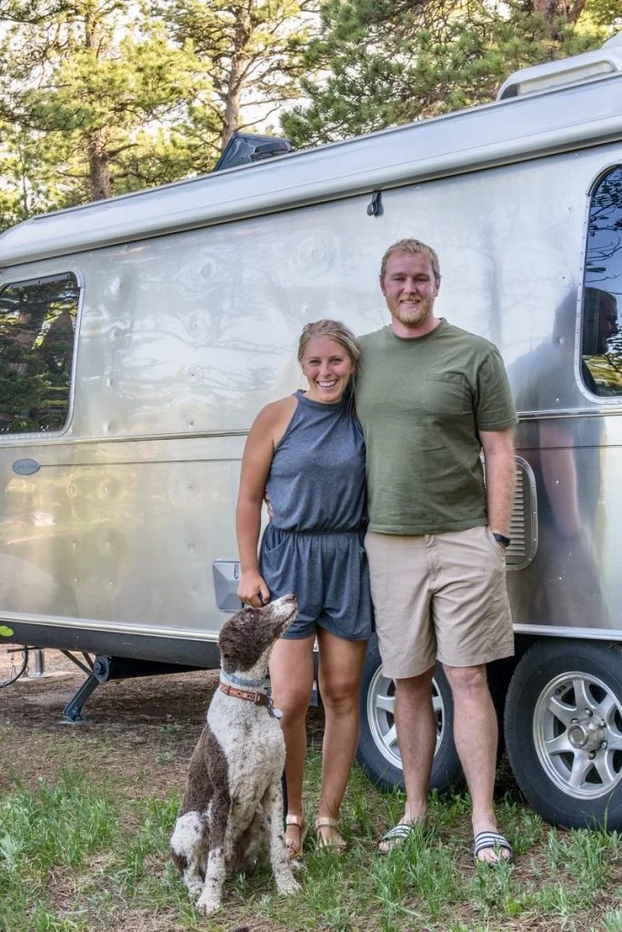 A smiling couple stands in front of a silver camper van surrounded by trees, with a brown and white dog sitting at their feet looking up at them.