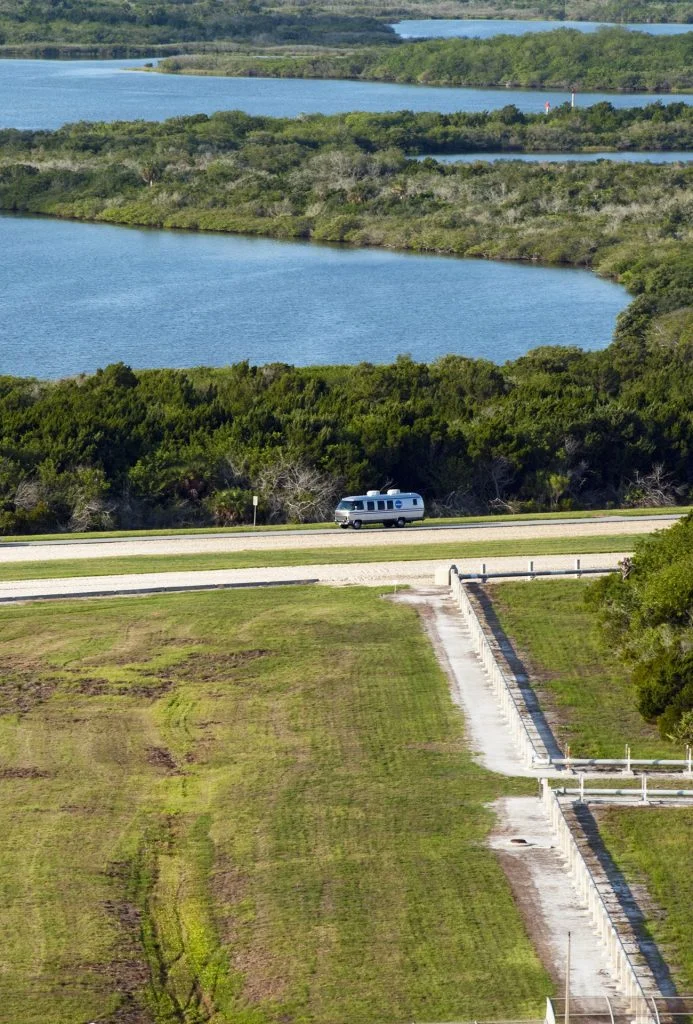 A lone white RV drives along a road bordered by grass and trees, with blue water and green vegetation in the background.