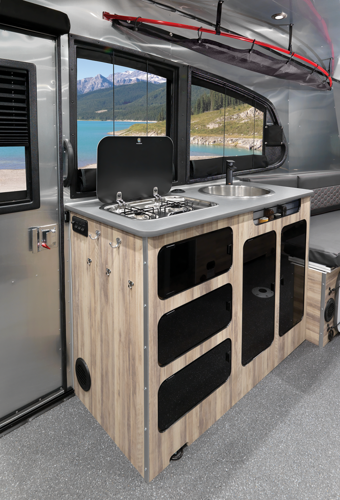 Compact RV kitchen with stove and sink, wooden cabinets, large windows displaying a scenic view of a lake and mountains outside.