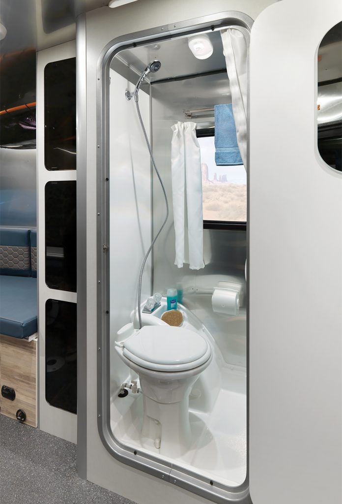 Compact RV bathroom with shower, toilet, and toiletries. Blue towel hangs on a rack, with window view of a desert landscape.