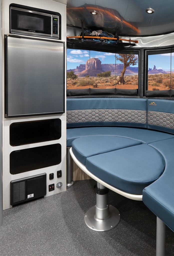 Interior of a modern camper with blue seating, a microwave, and a window view of a desert landscape with rock formations.