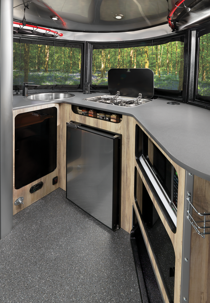 Compact camper van kitchen with sink, stove, microwave, and mini-fridge. Large windows reveal a lush forest view.