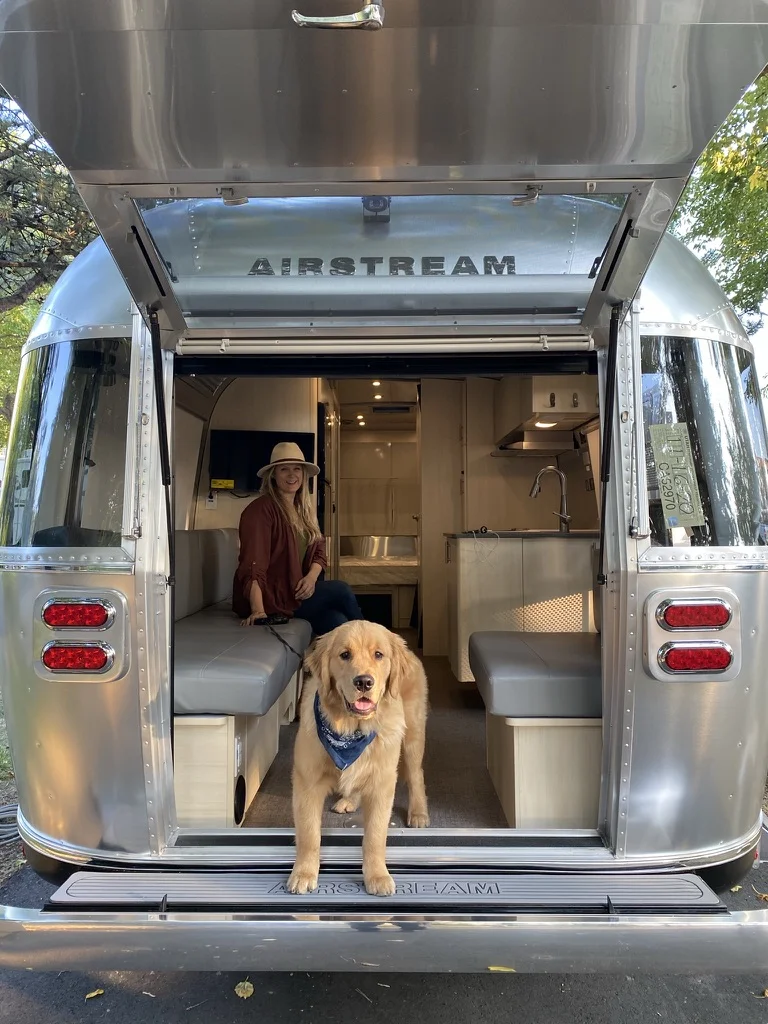 A woman in a hat sits inside an Airstream trailer while a golden retriever wearing a blue bandana stands at the open rear entrance, looking outside. The trailer interior appears modern and well-lit.