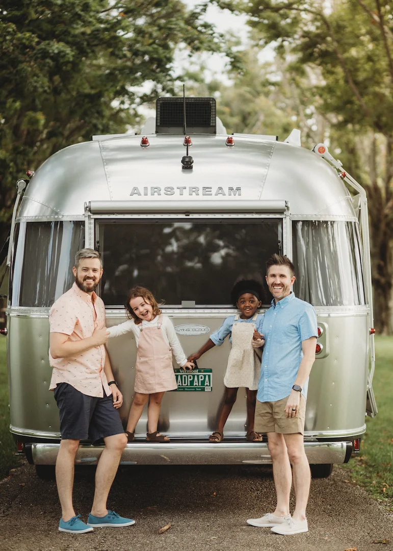 A family of four, two adults and two children, smiling and standing behind an Airstream trailer in a lush, green outdoor setting.