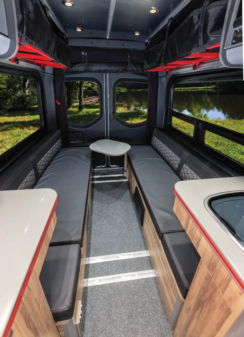 Interior of a modern camper van with a U-shaped seating area, small round table, and wood-accented storage. Large windows reveal a forest and lake view.
