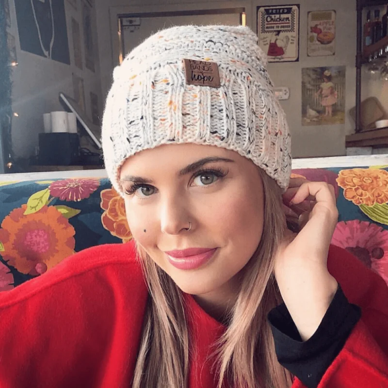 A woman with long blonde hair wears a knitted white beanie and a red top, sitting on a colorful floral couch in a cozy cafe with retro wall decor, smiling softly at the camera.