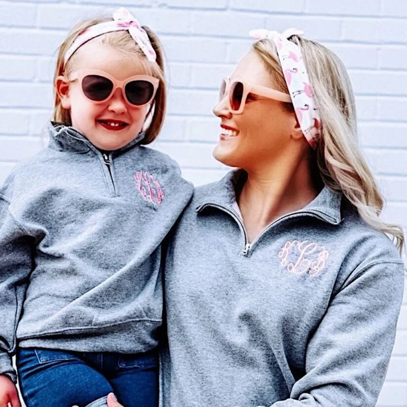 A woman and a young girl smile at each other while wearing matching gray zip-up sweaters, pink headbands, and pink sunglasses, standing in front of a white brick wall.