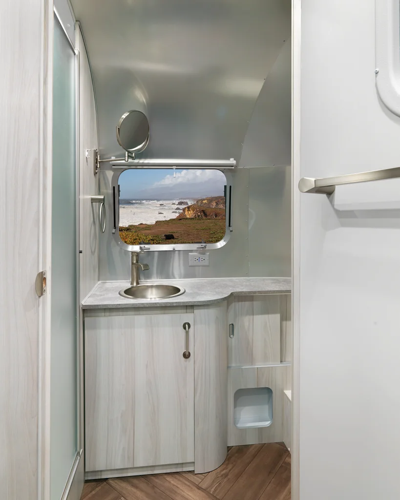 Modern RV bathroom with light wood cabinetry, a round sink, a small mirror, and a window above the counter. Through the window, an ocean view with waves and rocky cliffs can be seen.