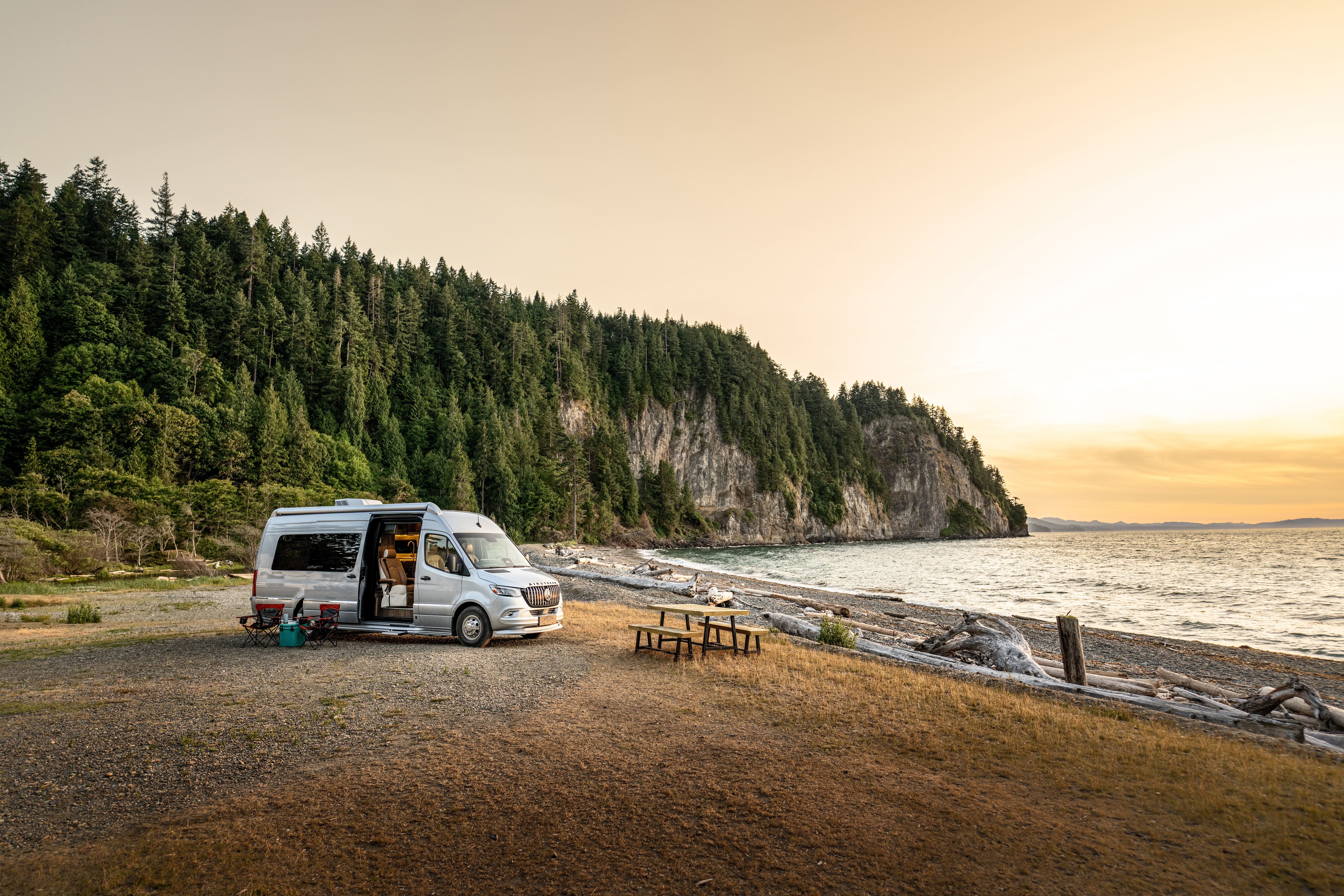 Camper van parked by a picnic table on a pebble beach at sunset, with a forested cliff and calm sea in the background.