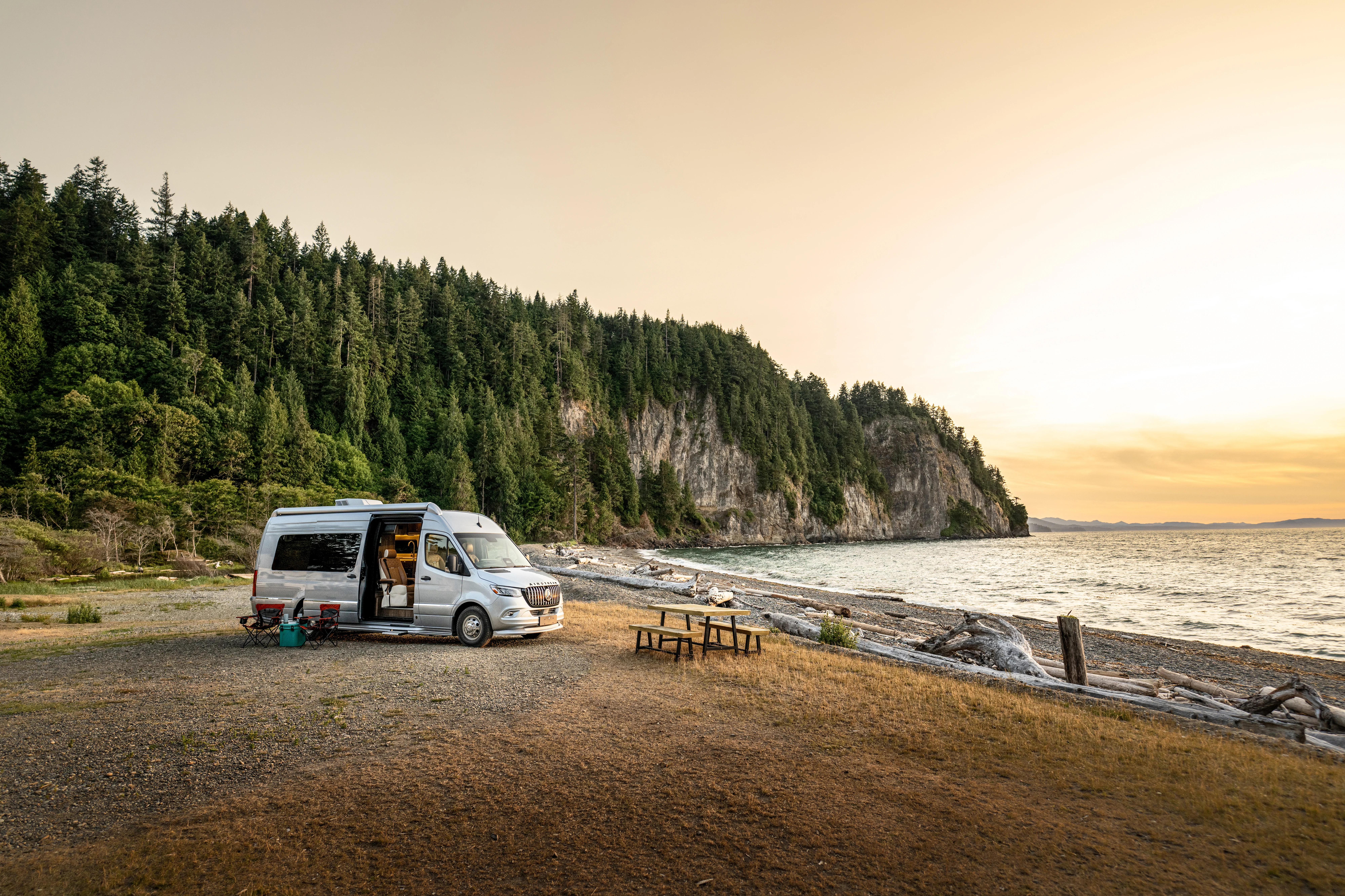 Camper van at a scenic coastal campsite with a rocky shore, picnic table, and forested cliffs under a golden sunset sky.