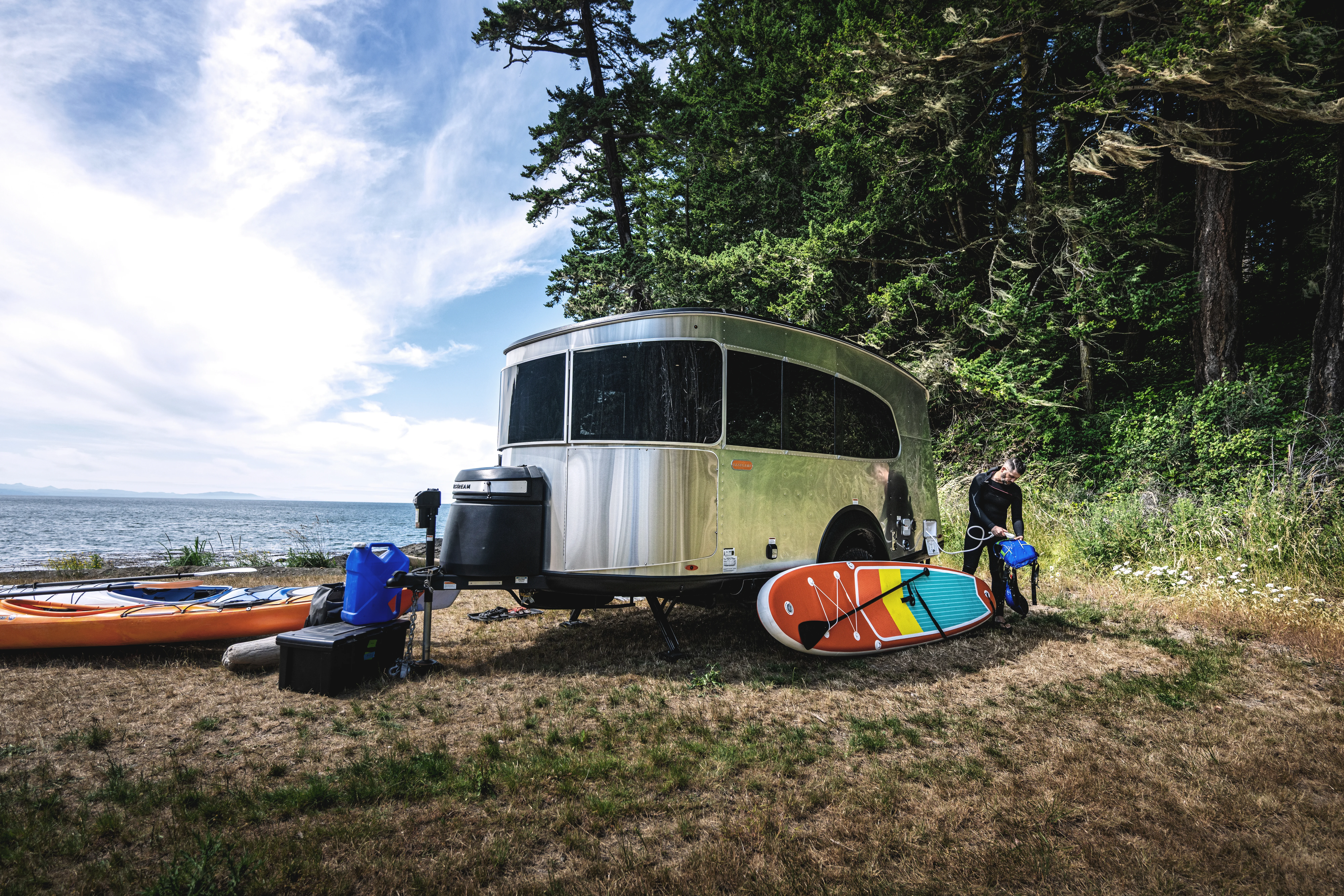 A silver trailer parked near a lake with kayaks and paddleboards nearby, surrounded by trees and grass under a partly cloudy sky.