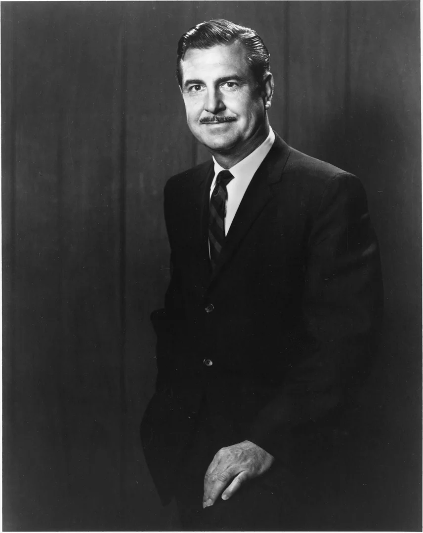 Black and white portrait of a man in a suit with a mustache, standing against a plain backdrop.