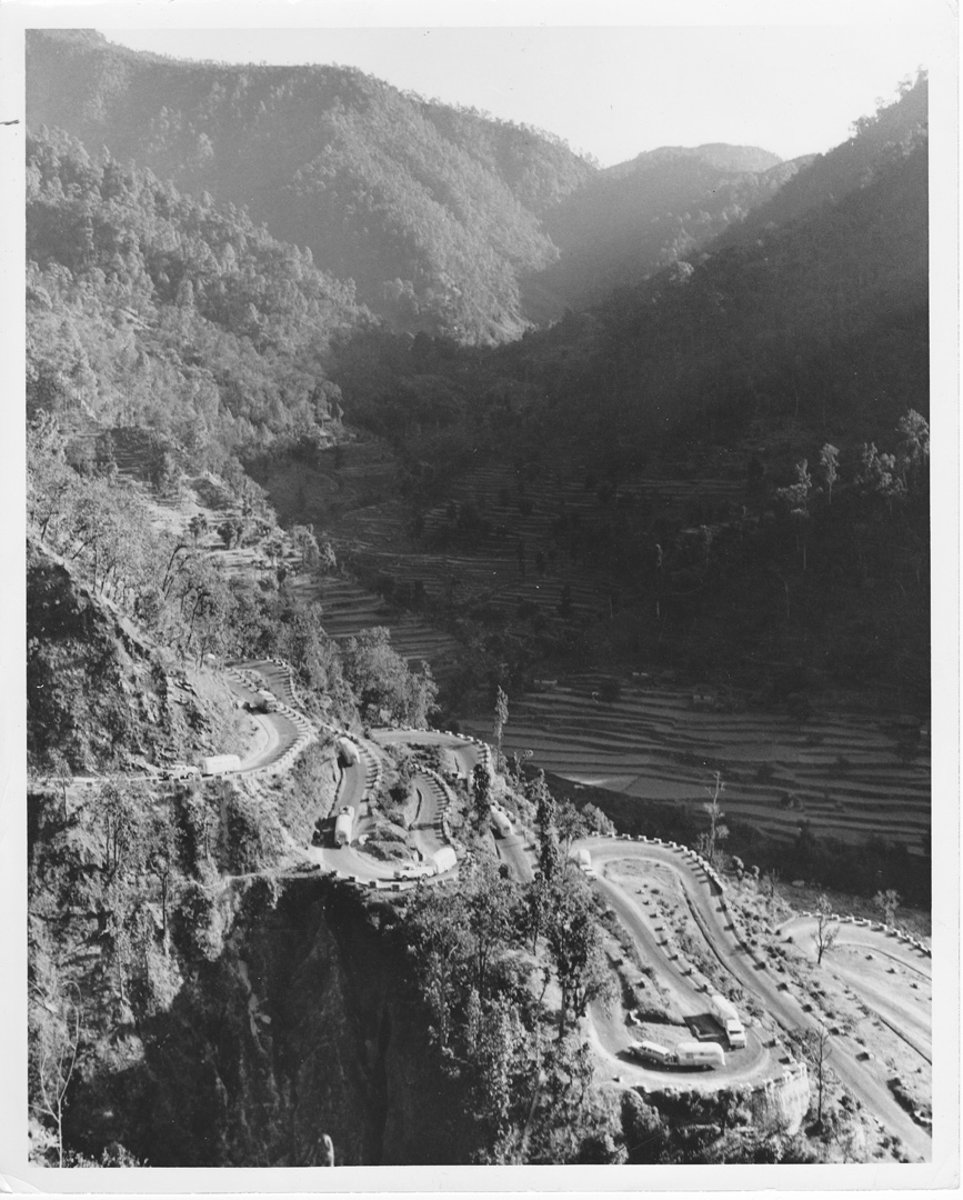 A winding mountain road with sharp hairpin turns passes through a lush, forested valley. Terraced fields are visible on the slopes, and several vehicles travel along the road. The area is surrounded by tall hills.