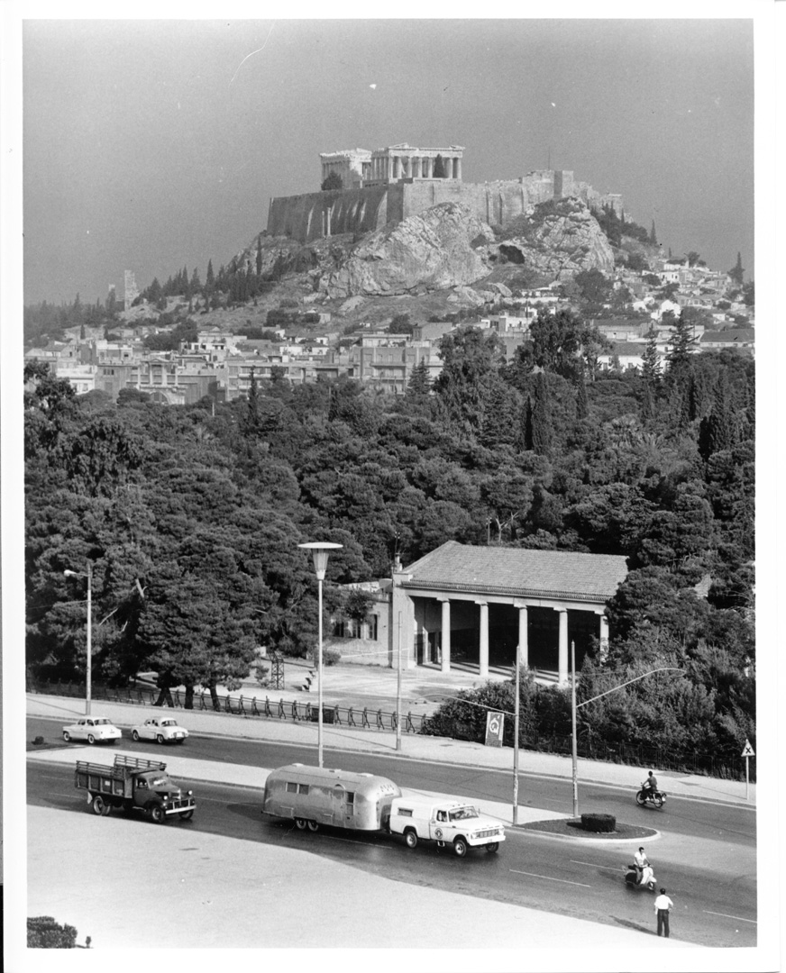 A black and white photo of the Acropolis on a hill above Athens, Greece. Below, cars, a trailer, a bus, and a motorbike travel a tree-lined street next to an ancient building with columns.