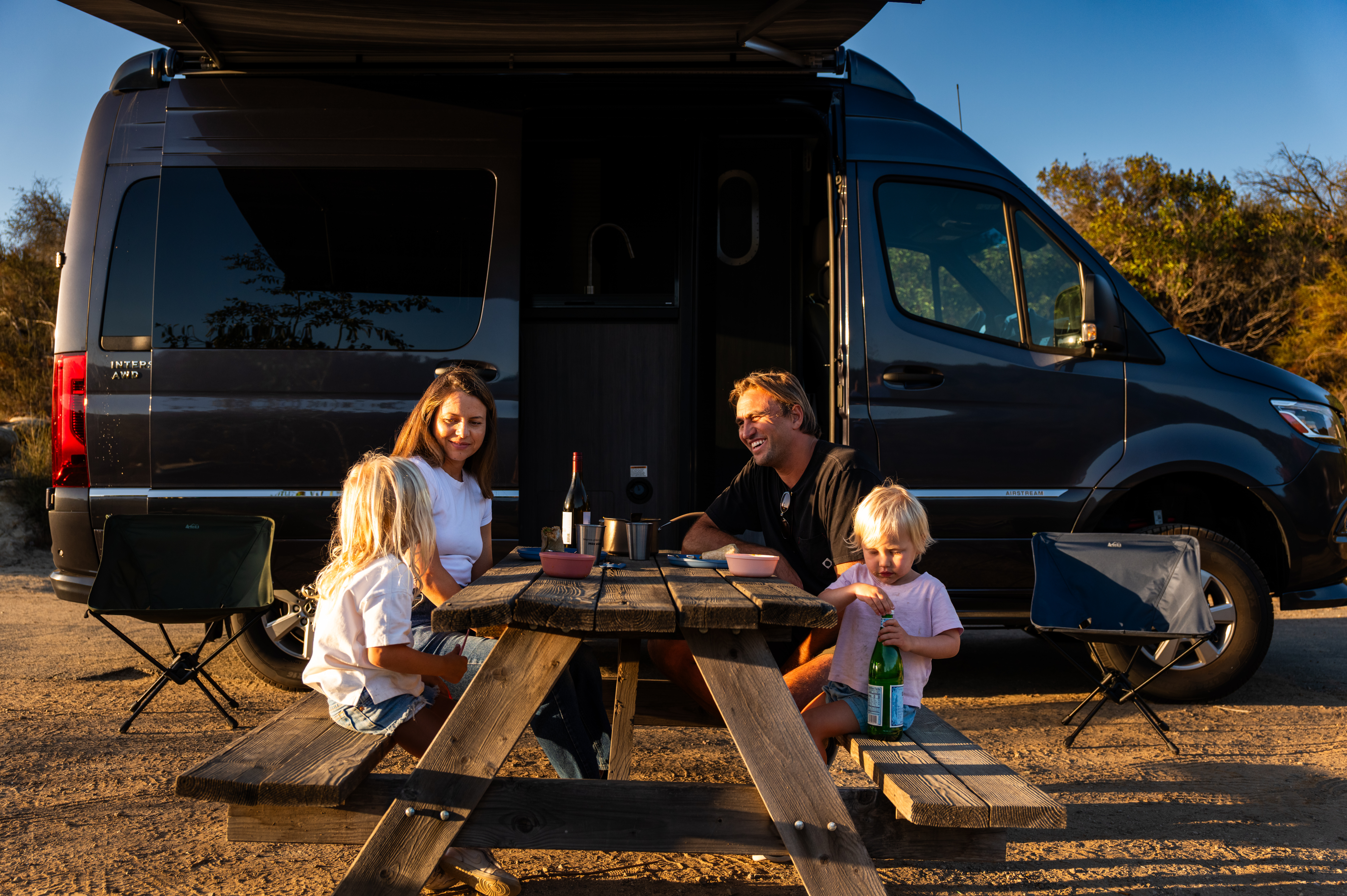 Four people gather around a picnic table beside an Airstream Interstate touring coach at an outdoor campsite.