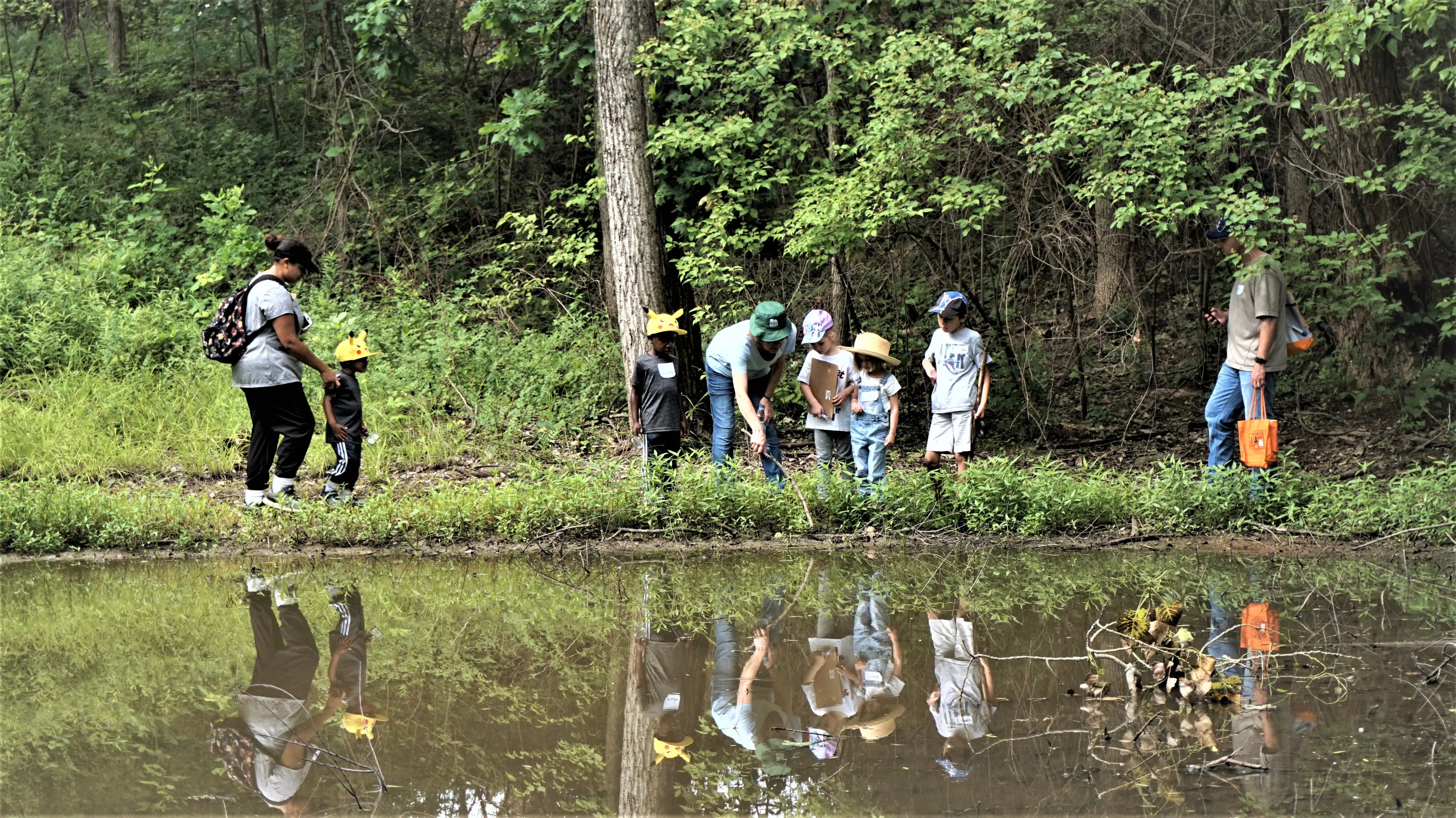 A group of people, including children wearing hats, explore a forest area near a pond, with their reflections visible in the water.