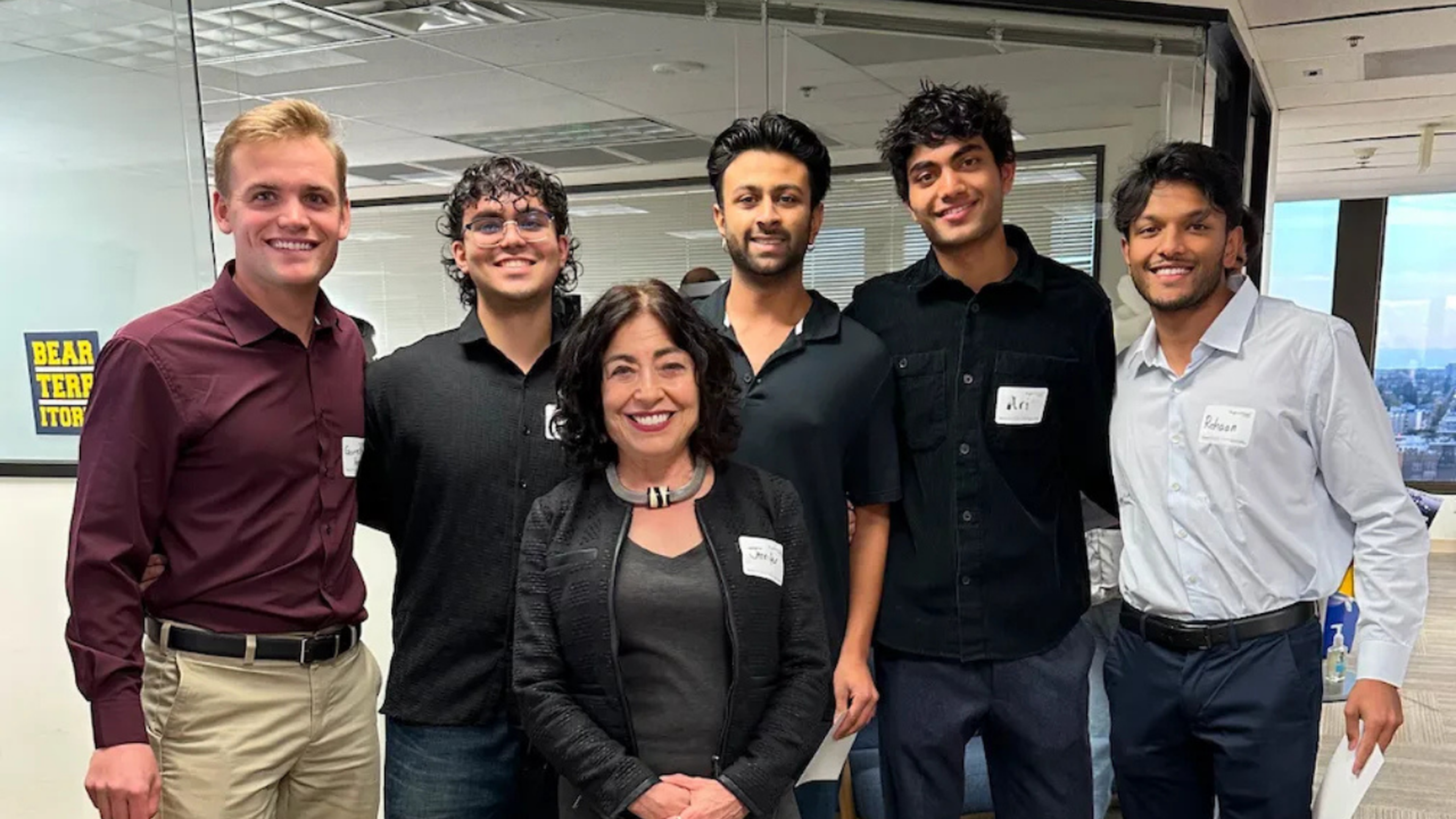 A group of six people smiling in an office setting, with name tags on their shirts, standing in front of a window with a city view.