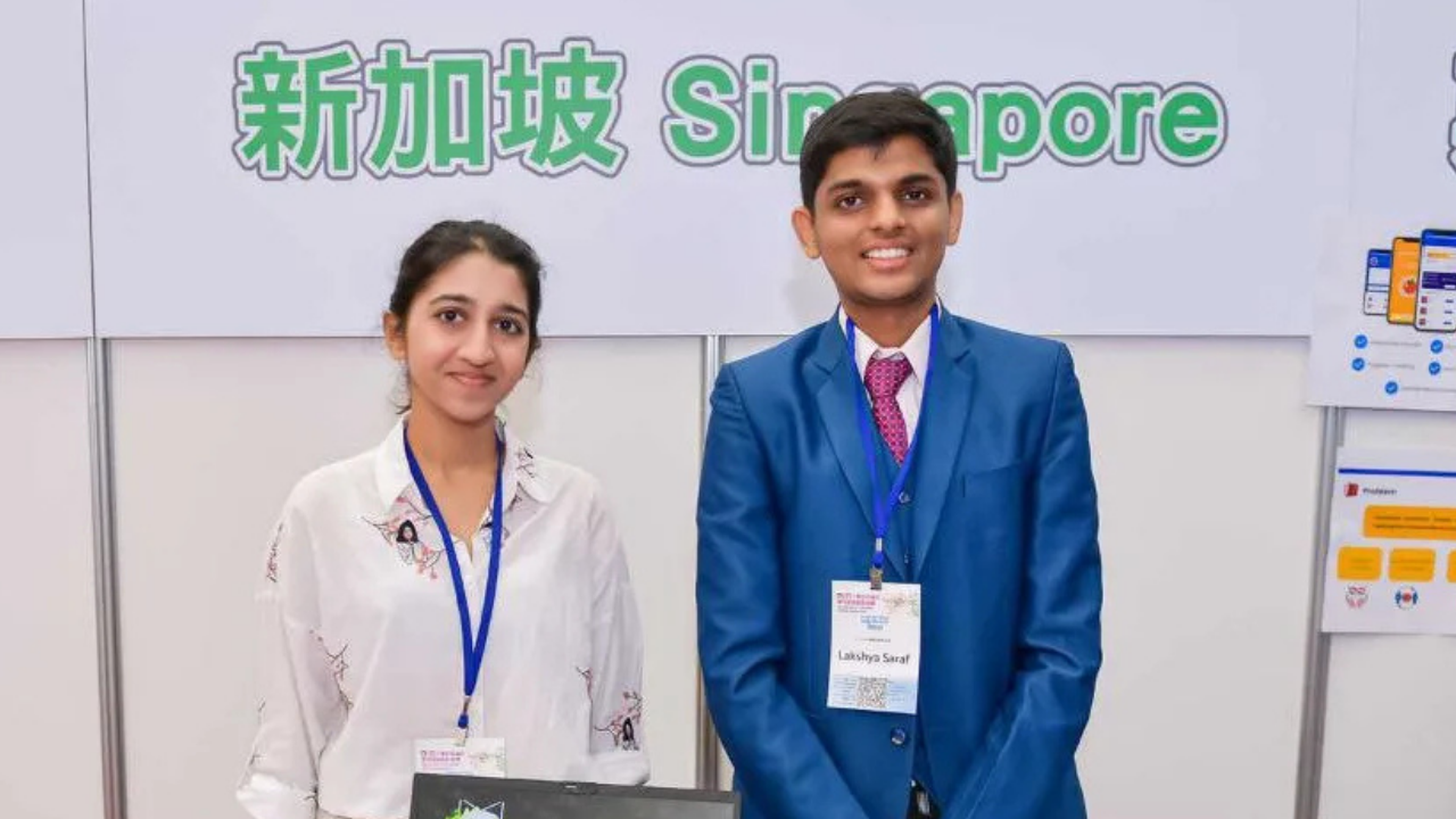 Two people stand smiling at a booth with a "Singapore" sign. They wear name tags and blue lanyards, with a laptop and display materials in front.