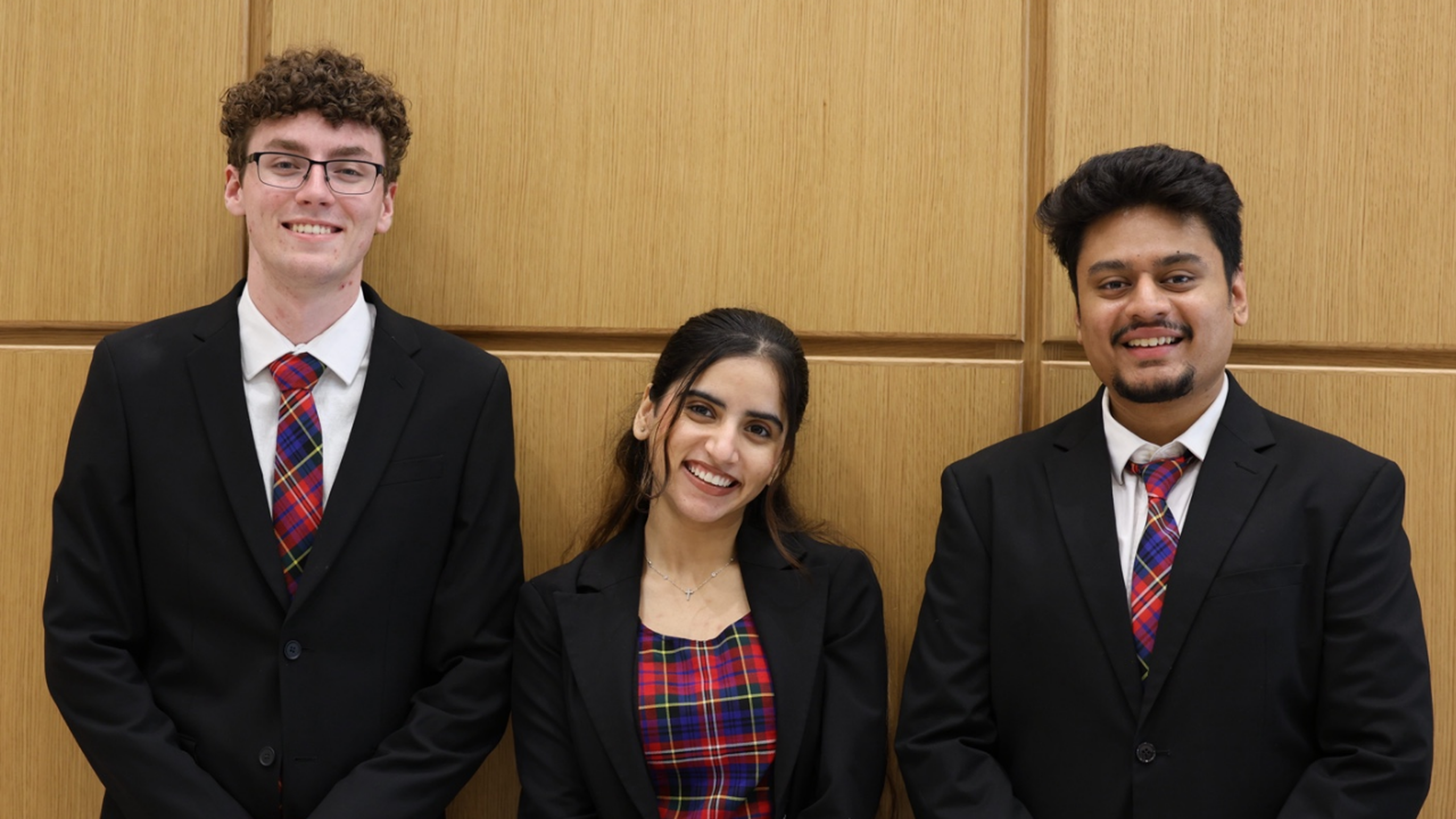 Three people in suits with plaid ties and shirts, smiling in front of a wooden panel background.