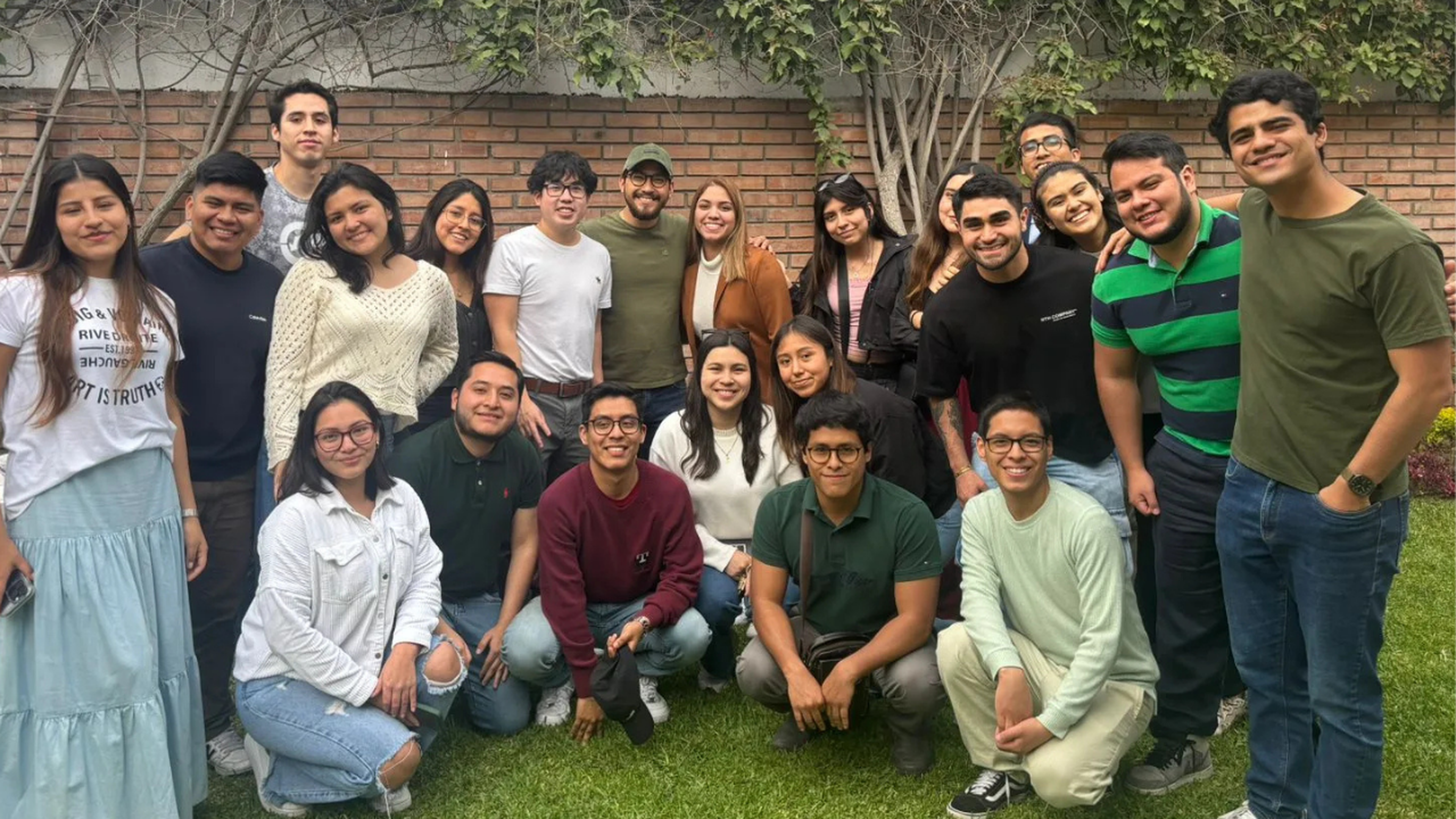 A group of people smiling and posing together on a lawn, with a brick wall and greenery in the background.