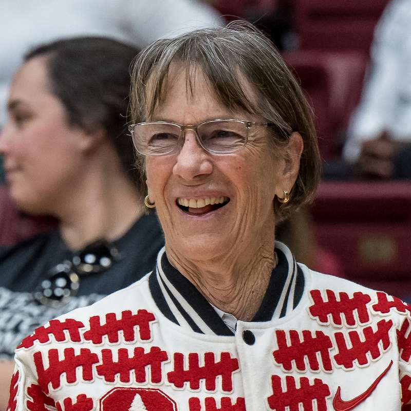 Stanford women’s basketball head coach Tara VanDerveer standing on the court sidelines, looking away from the camera and smiling genuinely.
