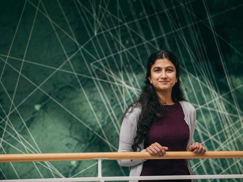 Aditi Sheshadri stands at a railing, smiling in front of a large green mural with intersecting white lines.