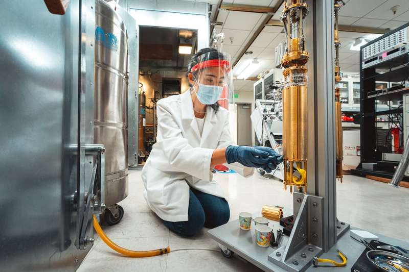 A researcher in a lab coat, face shield, mask, and blue gloves kneels to adjust a gold-colored quantum cryostat instrument in a physics lab.