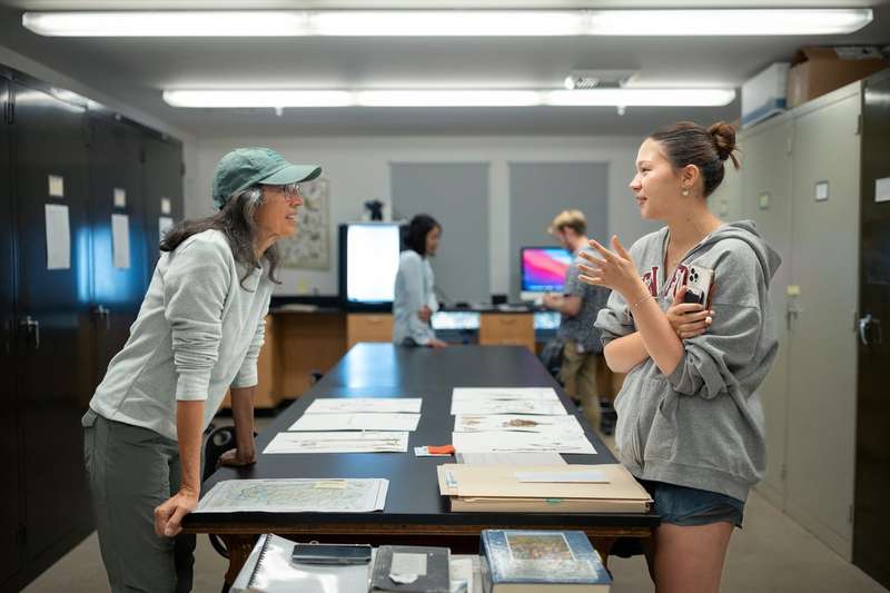 Two women talk across a long table covered with pressed plant specimens on paper in a herbarium lab, with other students working at computers behind them.