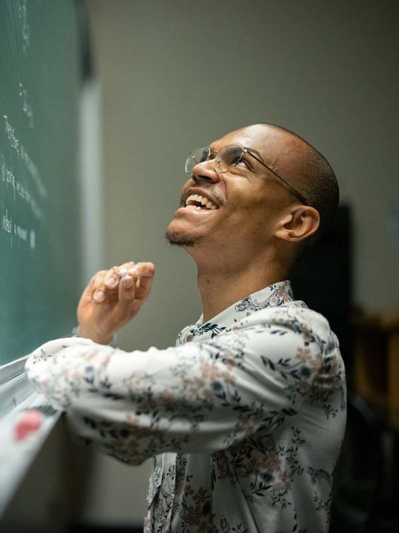 Jadon Geathers smiles and looks up at a chalkboard, holding chalk mid-thought while working through a problem in a classroom.