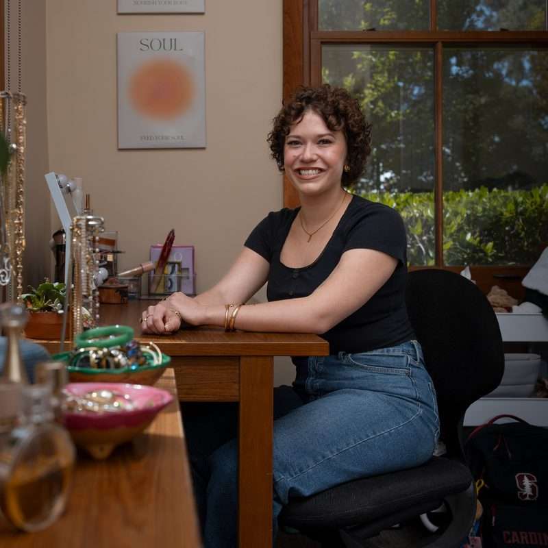 A college student sits at the desk in her dorm room, with jewelry and a plant in the foreground