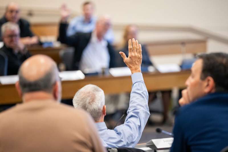 A person raises their hand to vote or ask a question during a faculty senate meeting, seated among others at tables in a wood-paneled hall.
