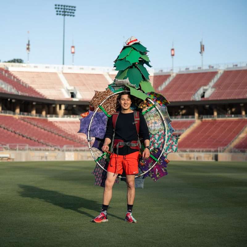 Grayson Armour stands smiling on an empty stadium field wearing an elaborate handmade Stanford Tree mascot costume built from a tree shape, hoop, and colorful umbrellas.