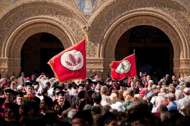 Two red Stanford tree-crest flags held high above a crowd of graduates in caps and gowns outside the carved stone arches of Stanford Memorial Church.