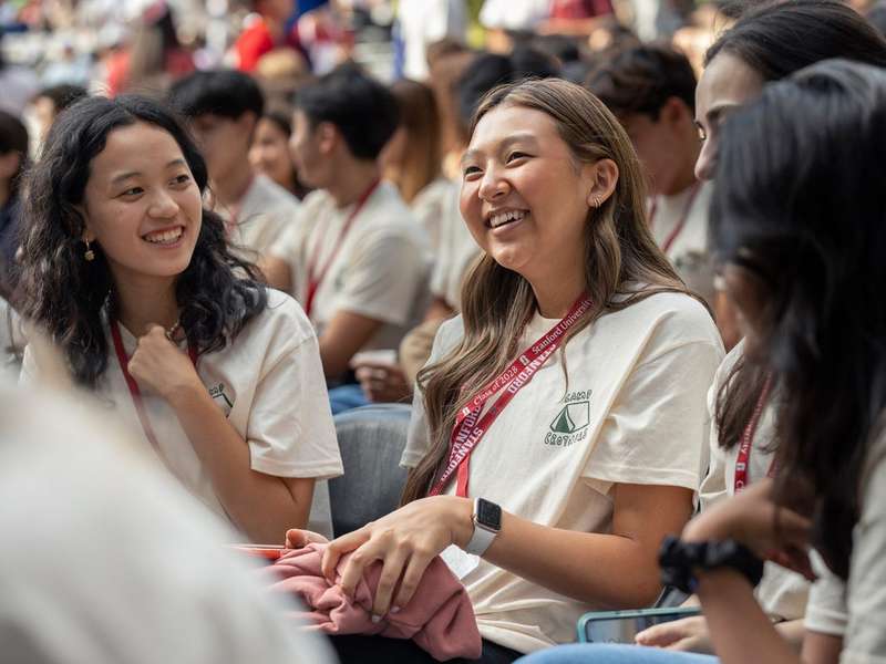 Smiling Stanford students in matching cream t-shirts and red lanyards chat together while seated in a crowd at a new-student convocation event.