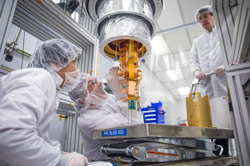Two scientists in white cleanroom suits and hairnets closely inspect a gold-colored scientific instrument suspended on a stand, while a third looks on, in a SLAC cleanroom.