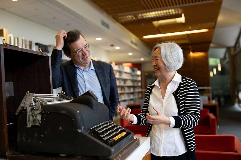 Two people laugh together in a library beside the MingKwai, a vintage Chinese typewriter with a large keyboard, displayed in an open wooden case.