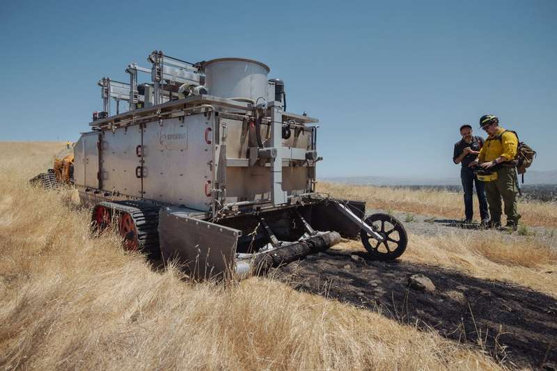 Burn bot leaves a trail of blackened grass amid California summer golden grass. Two men look on.