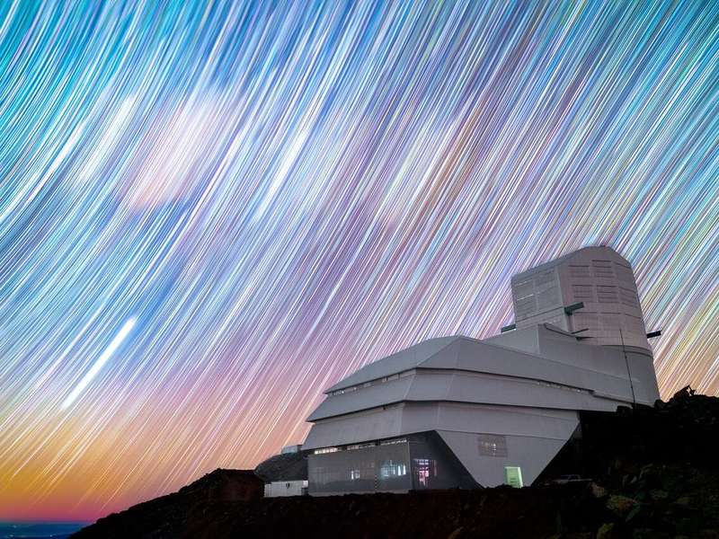 A timelapse shows the Rubin Observatory, a modern-looking angular building, standing in the foreground with trails of stars streaking in a colorful night sky behind