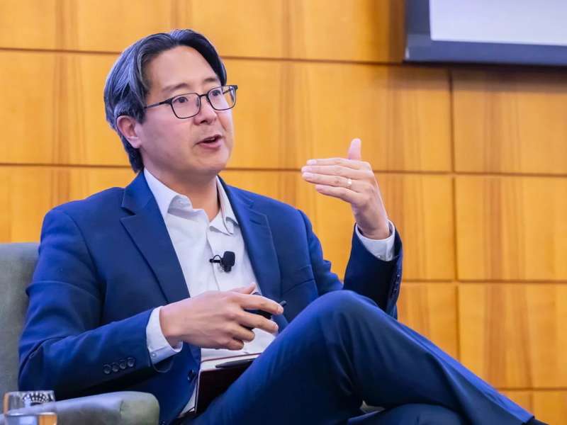 Michael Hsu in a suit speaks while seated in a chair, gesturing with one hand during a panel discussion.