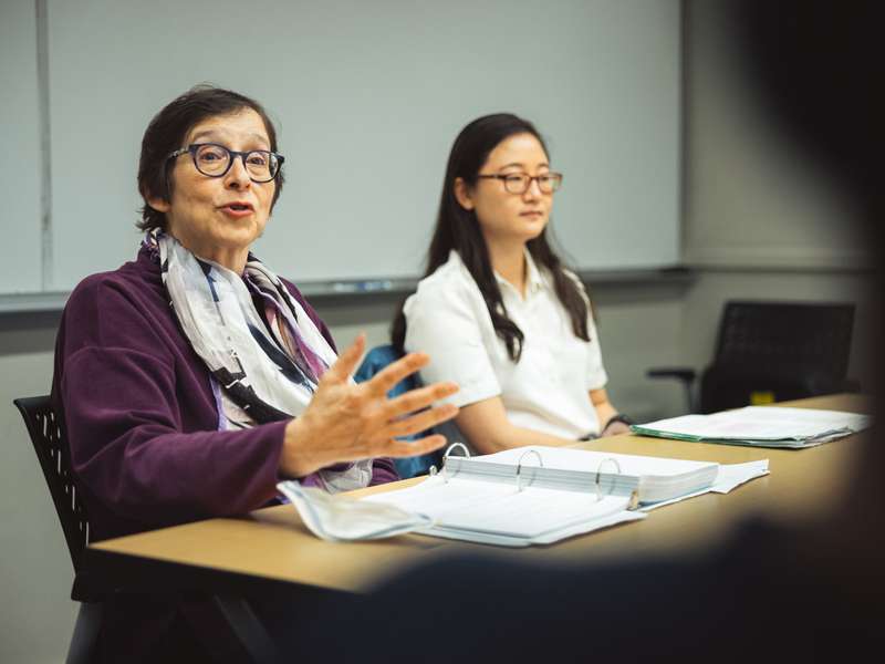 Pam Karlan gestures while speaking at a seminar table, with a student seated alongside her and open binders spread before them.