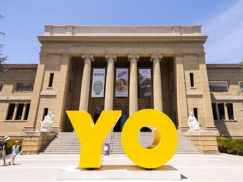 Two visitors walk past a giant yellow "YO" sculpture in front of the columned Cantor Arts Center, with exhibition banners hanging between the pillars.