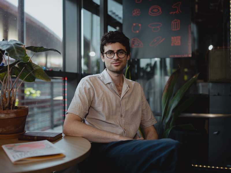 Vayu Hill-Maini sits at a lab cafe table beside a leafy potted plant, looking toward the camera.