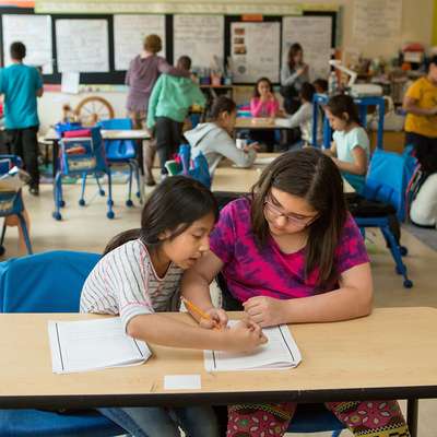 Two girls collaborate on assignments at a classroom table, while other students engage in various activities in the background.